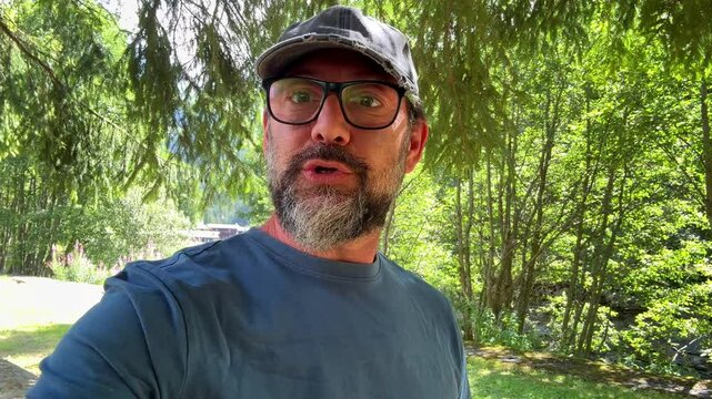 Male travel influencer with a beard, blue shirt, and cap walking in the mountains, engaging in a video call to share picturesque landscape views and his vacation experience with followers