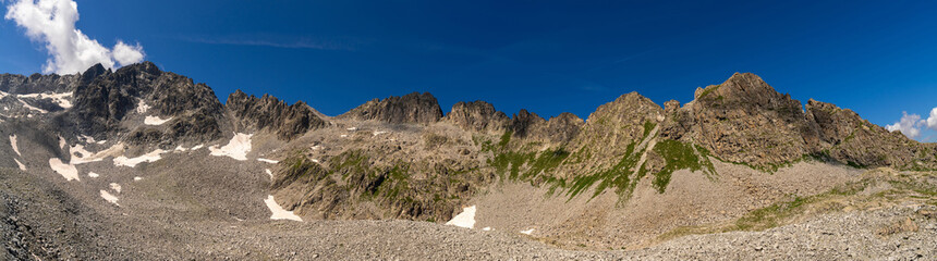 A dramatic panoramic view of rugged mountain peaks with patches of snow and expansive rocky terrain, set against a deep blue sky, showcasing the raw beauty of the highlands