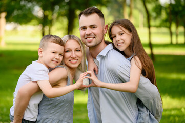 Fototapeta premium Beautiful parents with children pose for camera, holding boy and girl on their shoulders, spending time together in nature, showing unity, love. Happy parenthood,adoption, family , guardianship.