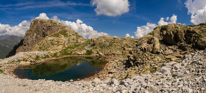 High altitude small alpine lake with clear reflections of rocky cliffs and rugged terrain, surrounded by pristine nature and panoramic mountain views under a bright blue sky