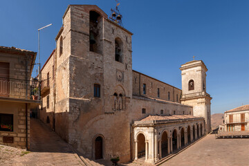 Panoramic view of Chiesa dei Santi Apostoli Pietro e Paolo in Petralia Soprana, Sicily, highlighting stone architecture, clock tower, and arched colonnade within village square under clear blue sky