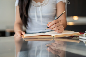 A close-up of a woman holding a pencil and writing in a book at a table indoors.