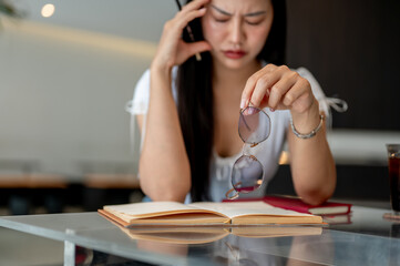 A close-up of a stressed woman takes off her eyeglasses to rest while reading in a coffee shop.