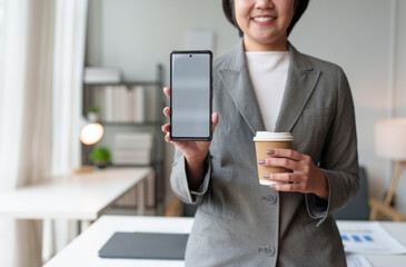 Asian woman, businesswoman sitting texting FaceTime or watching video on her smartphone while holding a cup of coffee drinking relaxing while working at home office. Work lifestyle concept.