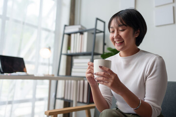 Asian woman, businesswoman sitting texting FaceTime or watching video on her smartphone while holding a cup of coffee drinking relaxing while working at home office. Work lifestyle concept.