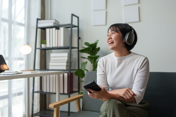 Bright Asian woman wearing headphones with mobile phone sitting, standing listening to music, dancing having fun and relaxing on vacation at home in living room. Concept of relaxation, lifestyle.