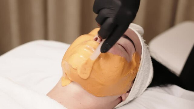 Close-up of a cosmetologist applying an alginate mask to a client's face with a spatula. The scene highlights professional facial skin care treatments in a beauty salon.