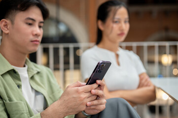 A serious Asian man is scrolling on his phone, ignoring his moody girlfriend.