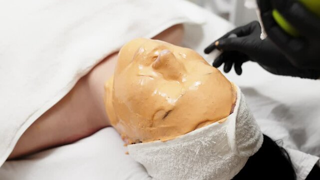 Close-up of a cosmetologist applying an alginate mask to a client's face with a spatula. The scene highlights professional facial skin care treatments in a beauty salon.