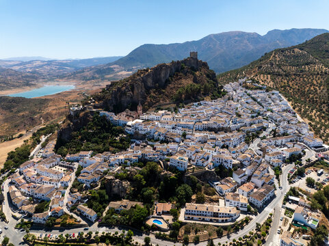 Aerial view of picturesque whitewashed buildings and historic castle on a hillside overlooking a serene lake, Zahara de la Sierra, Spain.