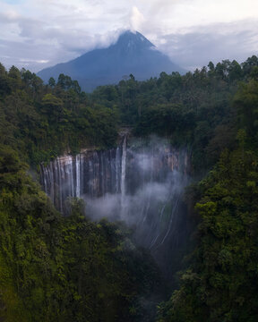Aerial view of a breathtaking waterfall with smoke rising from the majestic Merapi volcano, surrounded by lush forest, Tumpak Sewu, Java, Indonesia.