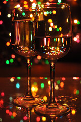 Two transparent wine glasses filled with water, with colorful Christmas lights on a tree in the background