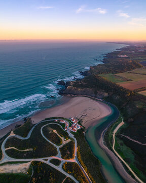 Aerial view of a picturesque beach at sunrise with a serene river and a tranquil village along the coastline, Odeceixe, Portugal.