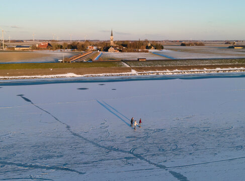 Aerial view of people ice skating on frozen ijsselmeer at sunset with a serene rural landscape and church, Cornwerd, Netherlands.