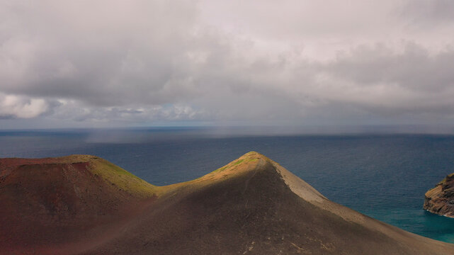Aerial view of a majestic volcanic landscape with a serene ocean backdrop and dramatic clouds, Capelo, Portugal.