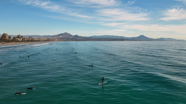 Aerial view of surfers riding waves along a beautiful coastal beach with mountains in the background, Alicante, Spain.
