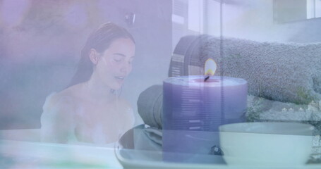 Image of scented candle and towels over caucasian woman in bath splashing face