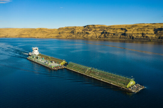 Aerial view of a river barge and tugboat navigating calm waters surrounded by serene hills under a blue sky, Columbia River Gorge, Benton County, United States.
