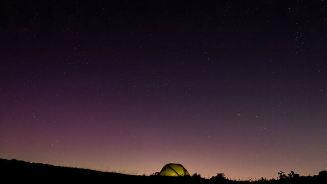 An illuminated wild camping tent on a hill summit under a starry night sky and a faint red tinge of the northern lights in the UK