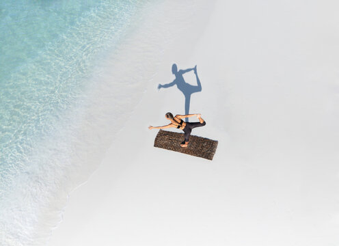 Aerial view of a serene woman practicing yoga on a turquoise beach with gentle waves and soft sand, Thoddoo, Maldives.