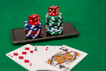 Close up of poker chips stacked on a smartphone with red dice on top and playing cards on a green felt background