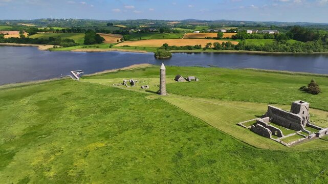 Devenish Island, County Fermanagh, Northern Ireland, June 2023. Drone orbit clockwise over monastery ruins and 12th century Round Tower with long shadow spread across the grassy fields.
