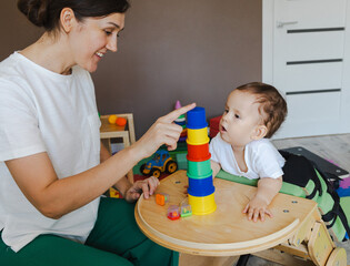 Cute little boy with hypotonia interacts with sensory therapist, focused on stacking colorful cups...