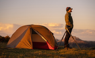 A man standing beside his wild camping tent and camera and tripod, on a hill in the countryside