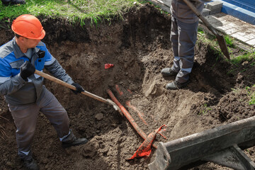 Laborers are seen carefully digging up red pipes buried in a dirt hole as part of an underground...