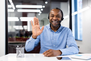 Happy businessman wearing headset waving during video call in modern office setting. Engaging in a virtual meeting using phone. Professional communication and digital connection concept.