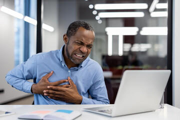 Obraz premium Mature businessman visibly in discomfort holding chest, sitting at desk with laptop open. Office ambiance with modern lighting. Scene capturing moment of unexpected pain during work setting.