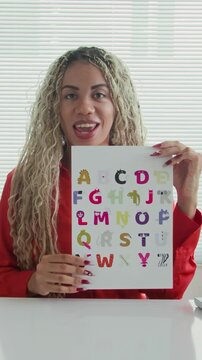 Vertical POV of female Hispanic logopedist talking on camera holding educational materials with alphabet at table
