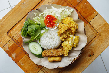 A plate of Indonesian Ayam Geprek fried chicken with white rice fresh vegetables and spicy chili paste is served on a rustic wooden table.
