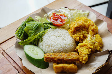 A plate of Indonesian Ayam Geprek fried chicken with white rice fresh vegetables and spicy chili paste is served on a rustic wooden table.