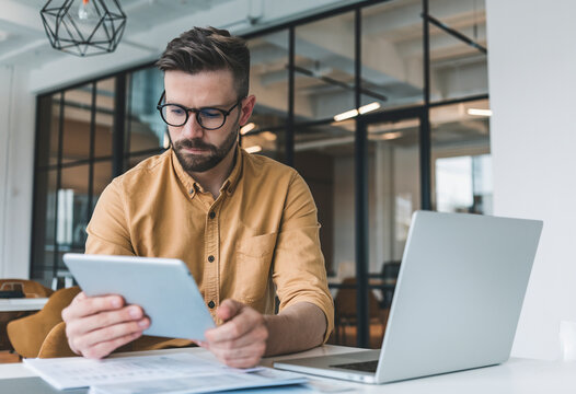 Serious businessman inside the office, utilizing a tablet and laptop to analyze data while reviewing online financial reports, showing deep concentration.