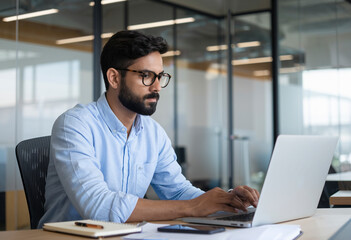 Serious Indian businessman inside his office, a mature man in a shirt and glasses, working diligently on his laptop while pondering a solution to a programming challenge.
