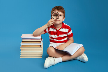 Serious smart kid in red striped shirt looking at camera through magnifying glass and holding open book while sitting crossed legged on floor near bunch of books, isolated over blue background