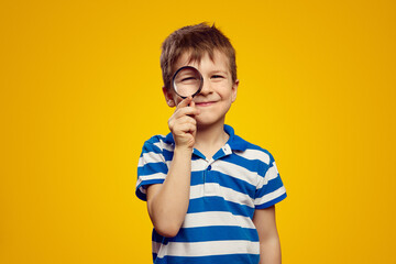 Curious funny kid examining environment with magnifying glass against yellow background, wearing...