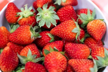 A portrait of a bunch of red delicious fresh strawberies with the green top in a white bowl. The tasty fruit is ready to be eaten.