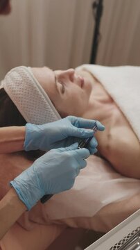 Aesthetician in blue gloves prepares a derma pen for a microneedling procedure on a female client in a spa setting.