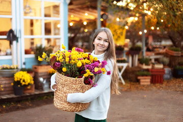 Cute smiling teen girl in a sweater, skirt  posing with bouquet of autumn flowers in basket. Young fashionable girl in knitted clothes with chrysanthemum flowers on backyard at home.