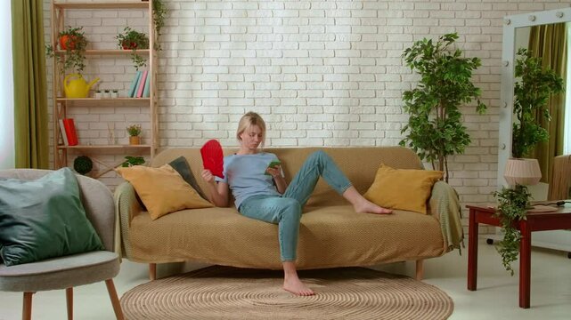 Young millennial woman sitting on the sofa in the living room waving fan coping with heat and texting on smartphone. Stuffiness everyday life concept.