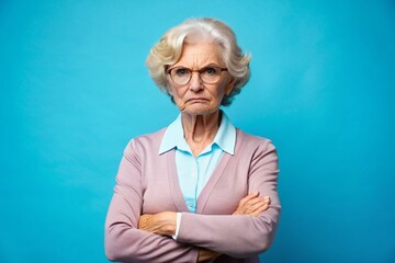 Scowling elderly educator with stern expression, arms crossed, stands against a bright blue backdrop, conveying frustration and discontent with her students' performance.