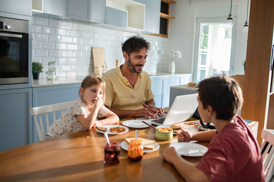 Father working on laptop while family has breakfast in kitchen - Powered by Adobe