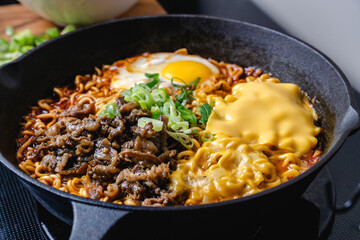 cooking savory ramen noodles topped with beef slice, egg and cheese in the cast iron skillet, with a spring onion as a garnish, kitchen setting