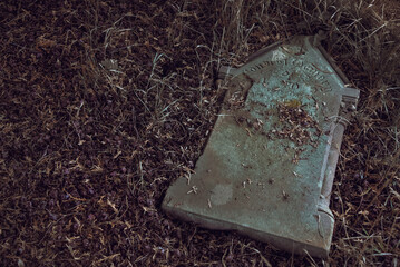 Spooky abandoned tombstone fallen on the ground of a cemetery. It seems like the ground is going to swallow de gravestone. Sensitive data has been remove or change.