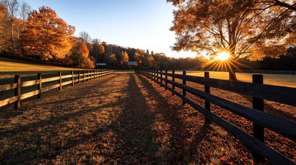 Obraz premium A picturesque scene of a farm in autumn with vibrant fall foliage, wooden fences, a grassy field covered with fallen leaves, and the sun setting behind a tree casting long shadows.