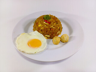 Plate of Indonesian nasi goreng (fried rice) served with a sunny side up egg and two chicken meatballs, neatly arranged on a white round plate against a clean white background