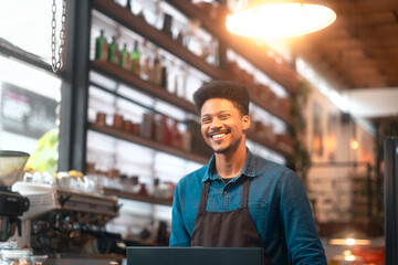 Fototapeta premium Friendly barista in a brown apron behind the café counter with a big smile. Cheerful coffee shop worker ready to serve customers with a warm and inviting demeanor.