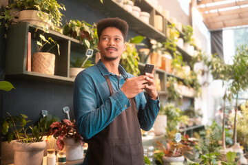 Optimistic florist in a brown apron using a smartphone while working in a flower shop. Smiling...